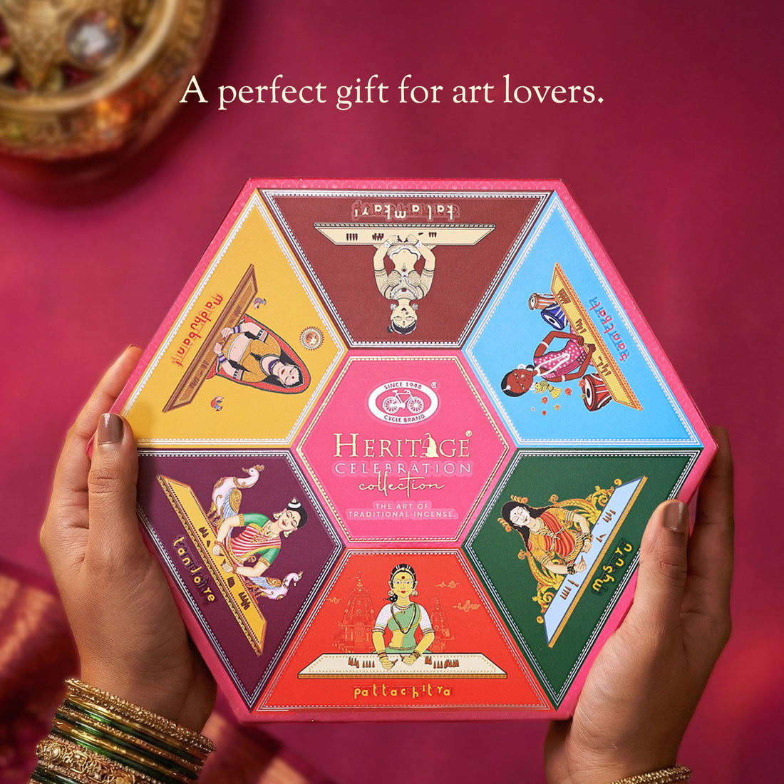 Hexagonal gift box with colorful designs held by hands on a pink background, labeled 'Heritage Celebration Collection'.